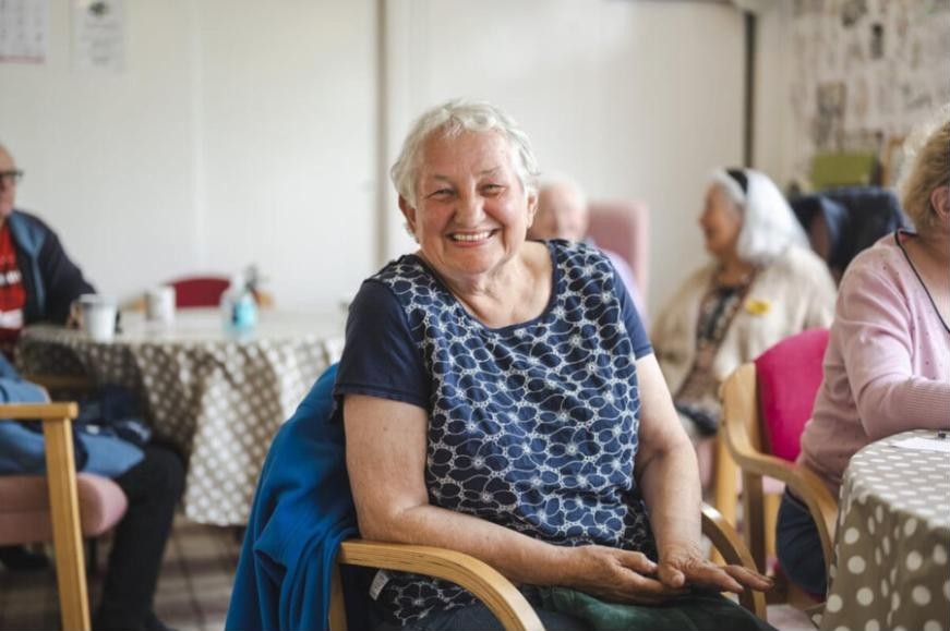 A smiling older woman sitting at a table with friends at a community group