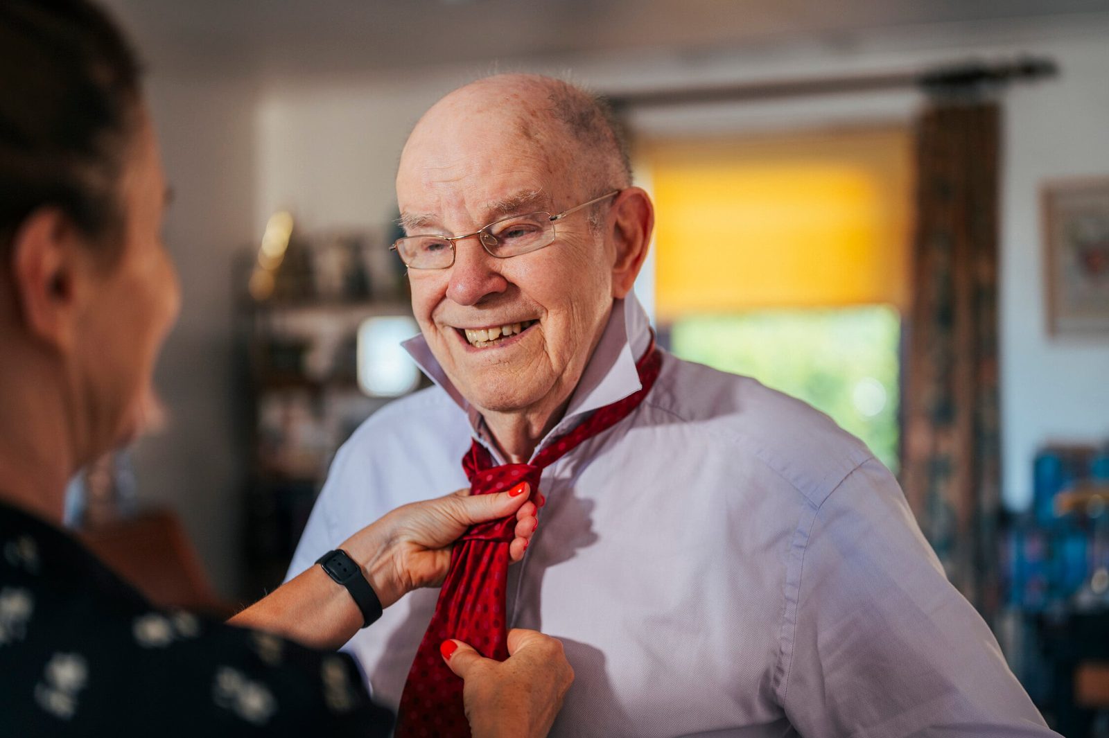 A carer helping a smiling older man with his shirt collar and tie at home