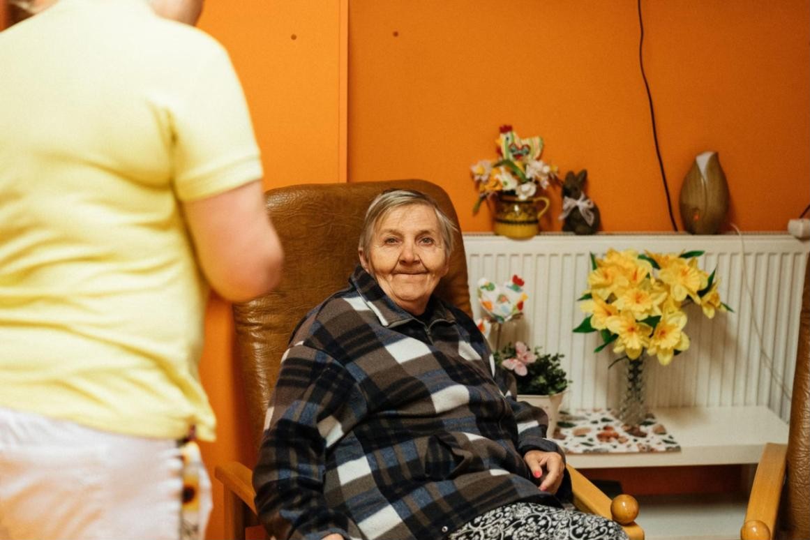A carer in a yellow uniform with a smiling older woman sitting in her armchair